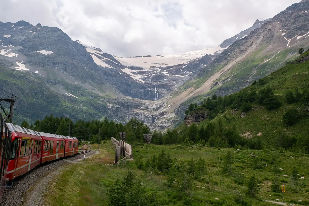 A red Bernina Express train curving through a green alpine valley, with the Palü glacier and waterfalls on the mountain ahead