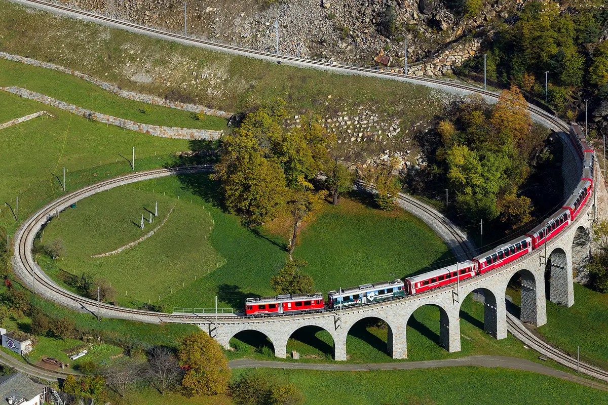 A red train crossing a curved stone viaduct that loops in a full circle over green fields, seen from above