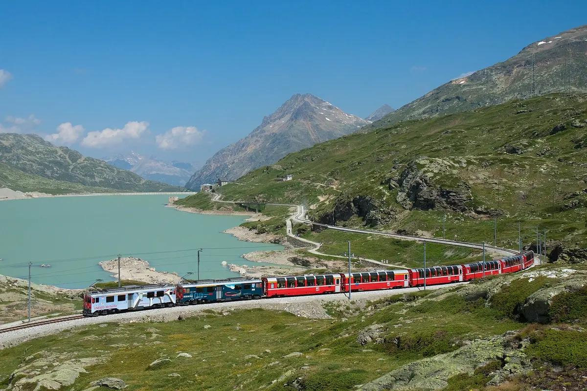 A red Bernina Express train with glass-roofed panoramic cars curving along the shore of a pale turquoise lake, with mountains behind